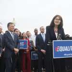 Mara Keisling stands at a podium with a crowd gathered behind her.