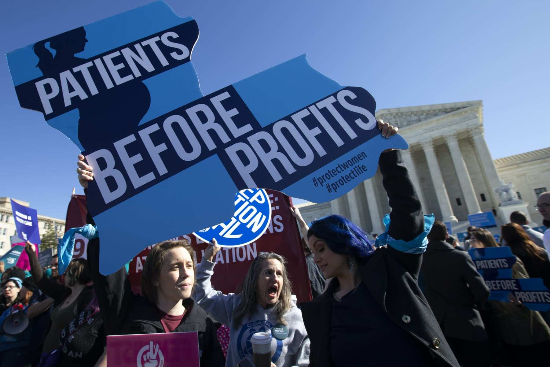 Protesters gather outside of the Supreme Court. The sign in the foreground reads "Patients Before Profits" and is shaped like Louisiana.