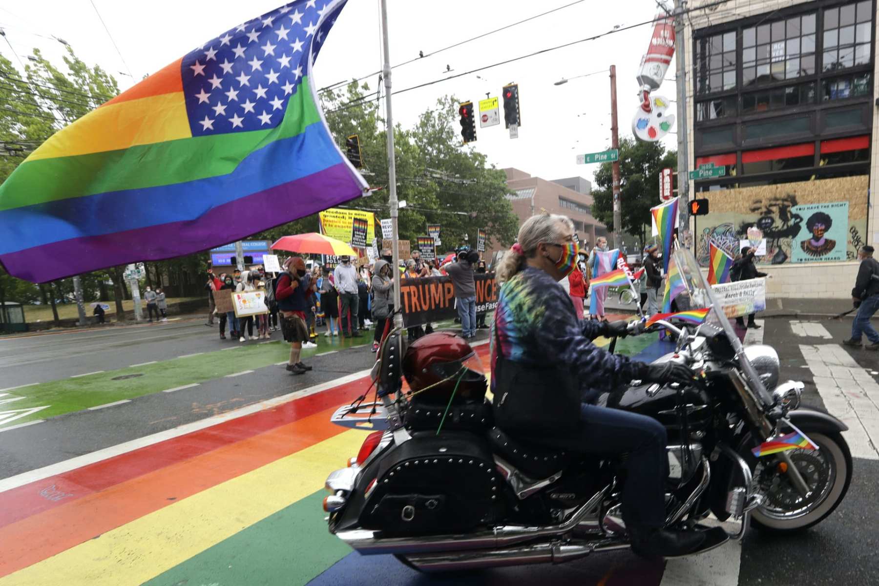 A person on a motorcycle with a rainbow flag attached to the back drives by protesters with a Trump-Pence sign.