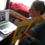 A teacher holding a book in front of the a virtual class.