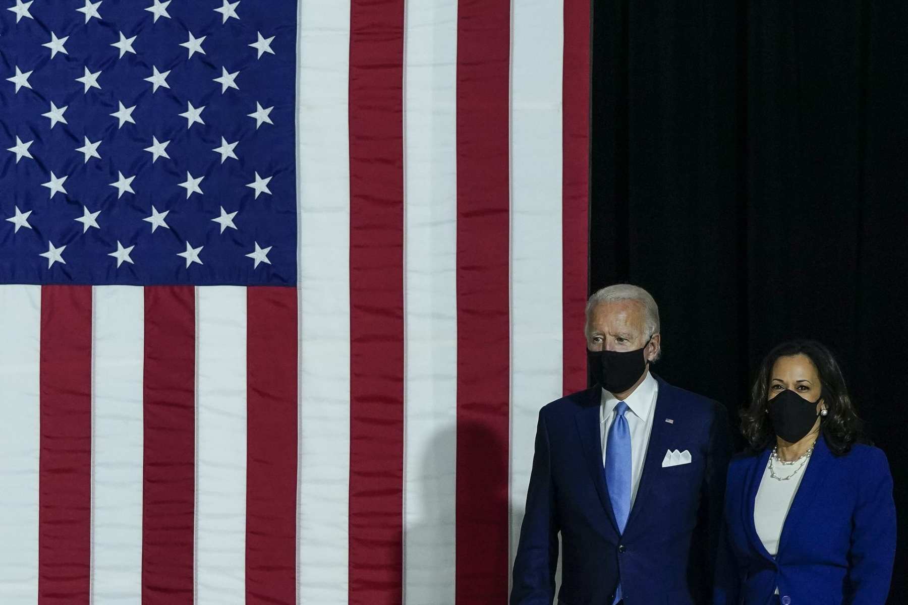 Joe Biden and Kamala Harris, both wearing masks, walk in front of an American flag.