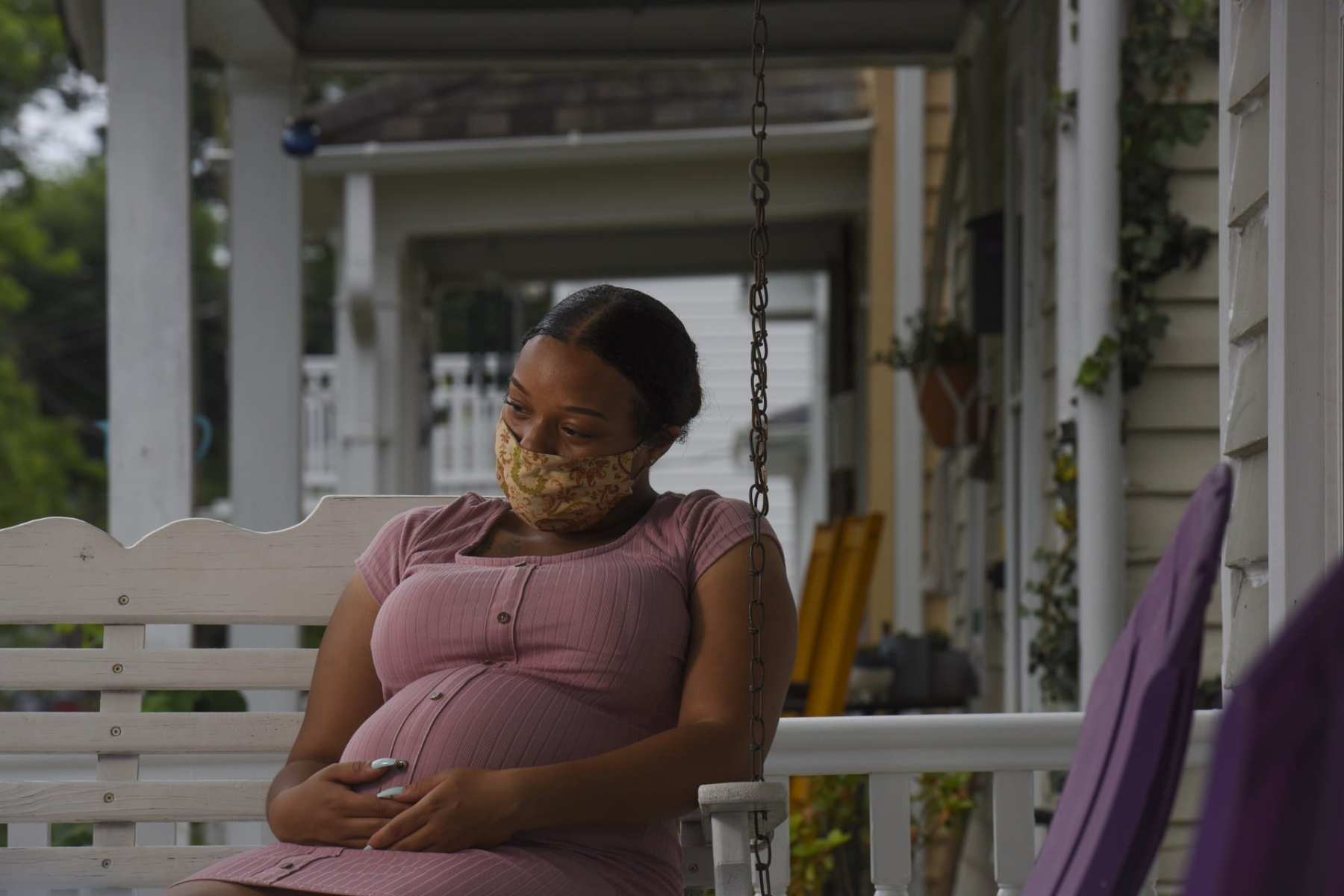 A pregnant woman sits on a porch swing holding her belly with a mask on.