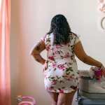 A woman in a floral dress stands with her back facing the camera with her hand on a crib.