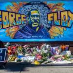 A woman holding a Black Lives Matter sign sitting before a mural of George Floyd, which is serving as a makeshift memorial.