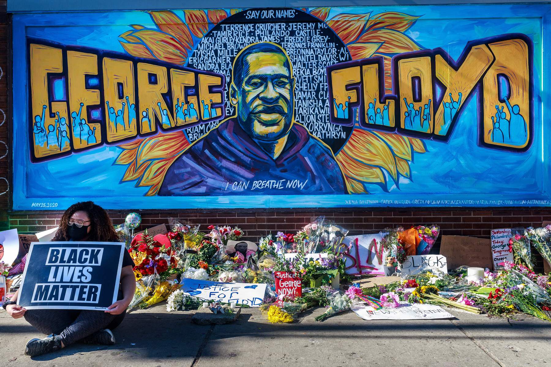 A woman holding a Black Lives Matter sign sitting before a mural of George Floyd, which is serving as a makeshift memorial.