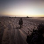 U.S. soldiers kneel in the sand looking at tanks in the distance at sunset.