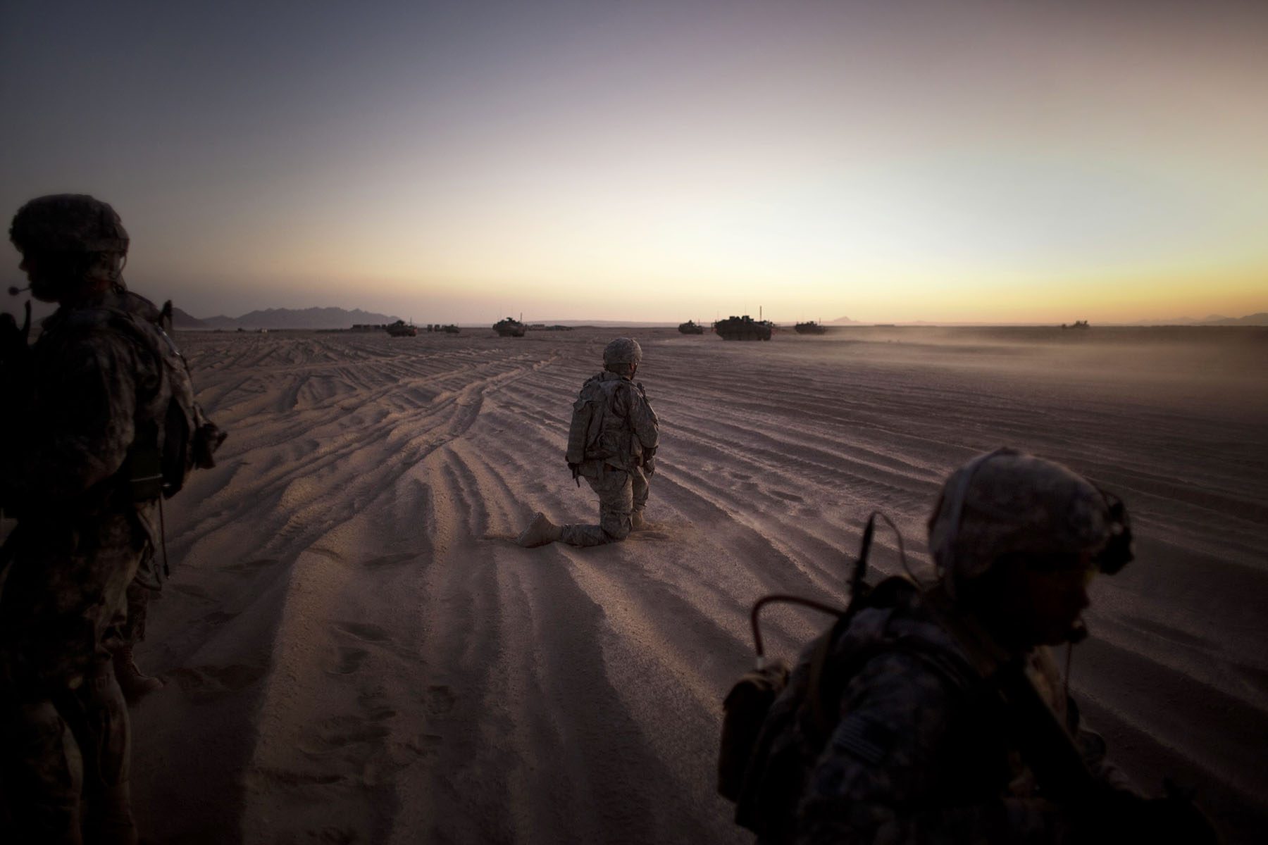 U.S. soldiers kneel in the sand looking at tanks in the distance at sunset.