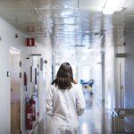 A doctor wearing a white coat is seen walking through a hospital corridor from the back.