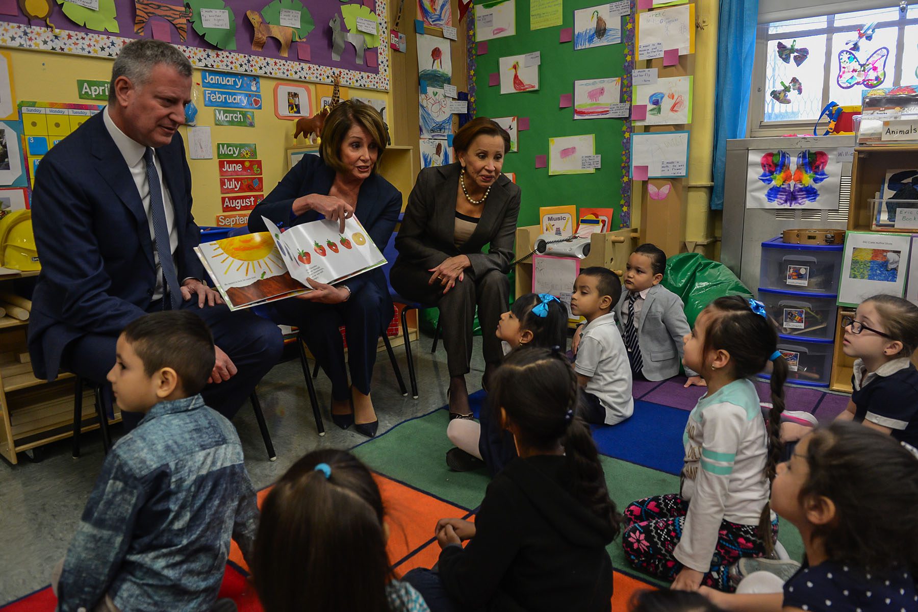 Nancy Pelosi, flanked by Bill DeBlasio and Nydia Velazquez points to a drawing of a strawberry in a children's book while children look on in a colorful classroom.