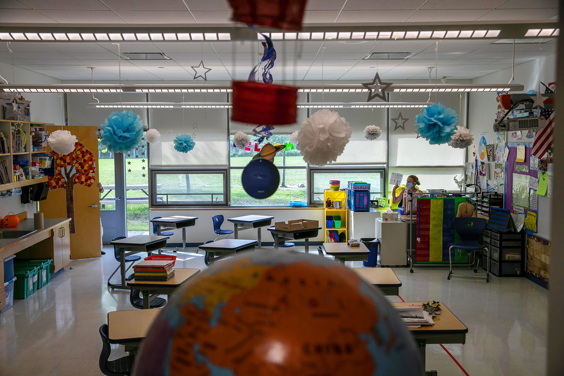 An empty classroom decorated with drawings of planets and trees is seen. A teacher is seen sitting at a desk and wearing a face mask.
