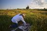 A man wearing a cowboy hat kneels to touch a tomstone in a field at golden hour.