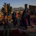 Colorful carpets are stewn about on the ground at an open air market while merchants sit, waiting for customers. Two women walk through, one of them carrying a small child. Their garments cover their face and body.