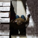 A man sits between two tables on which a laid stacks of blank ballots.