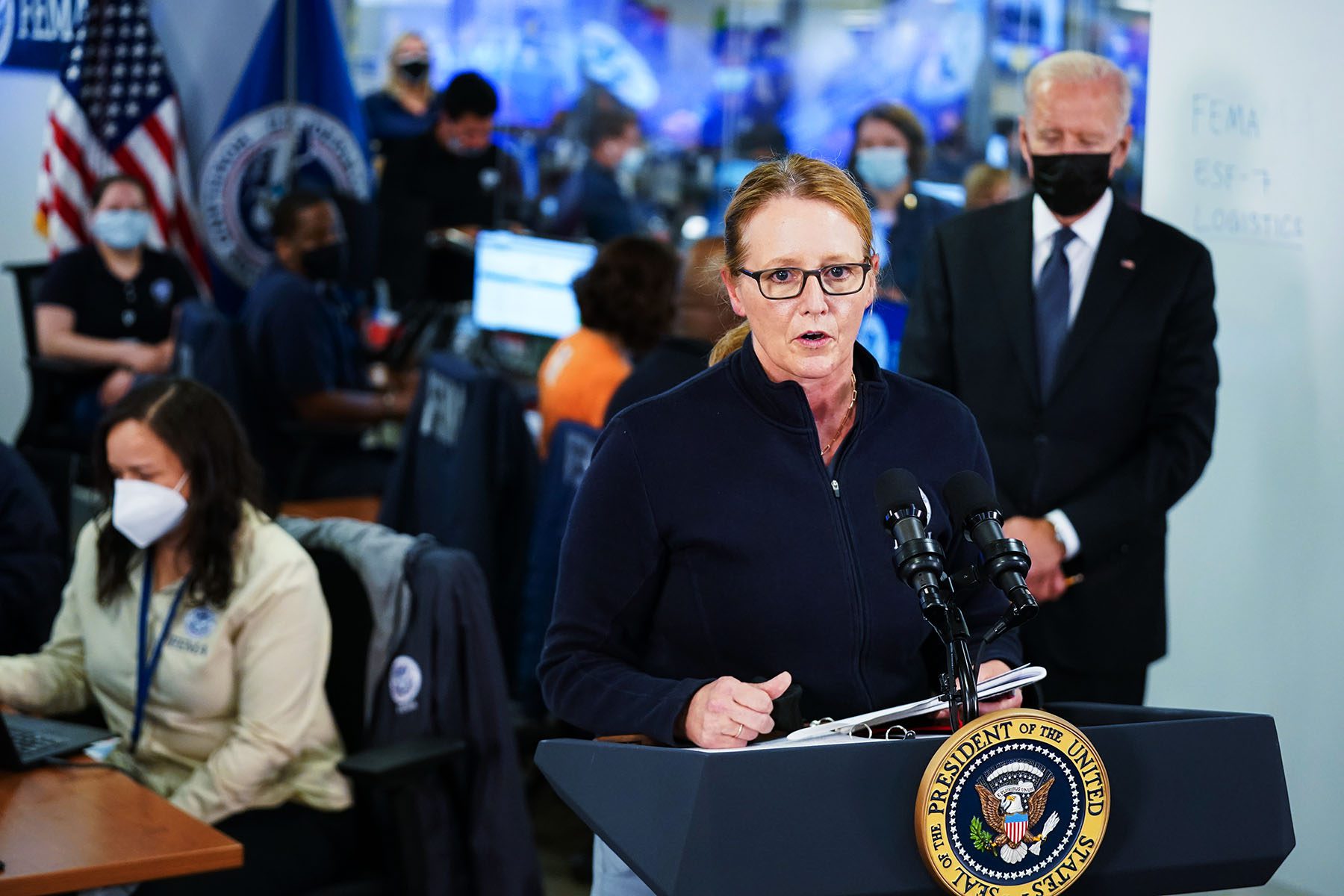 Deanne Criswell speaks at a podium on which is the presidential seal is hung. Joe Biden can be seen listening behind her.