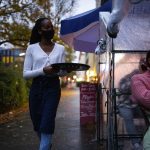 A young black waitress serves drinks to customers sitting in outdoor plastic tents.