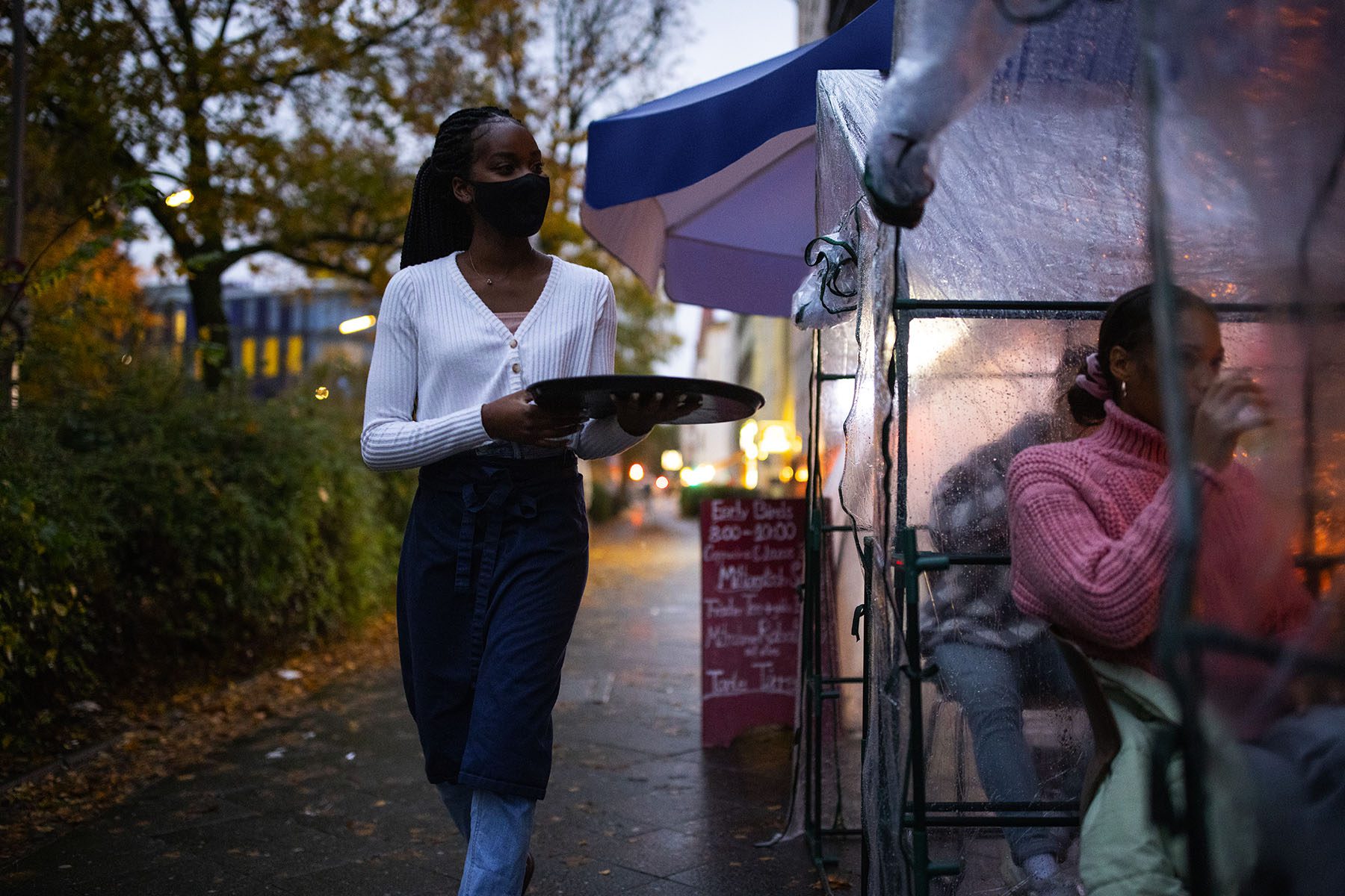 A young black waitress serves drinks to customers sitting in outdoor plastic tents.