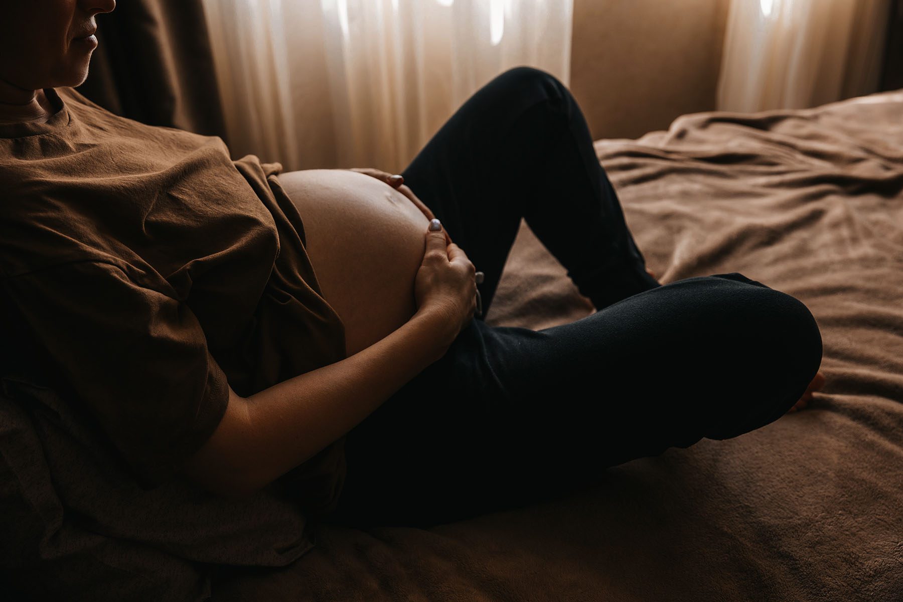 A pregnant woman holds her belly while sitting on a bed. Gold light floods the room.