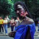 A participant wearing a golden necklace, earrings and a blue fabric as a top holds a flower to their cheek.