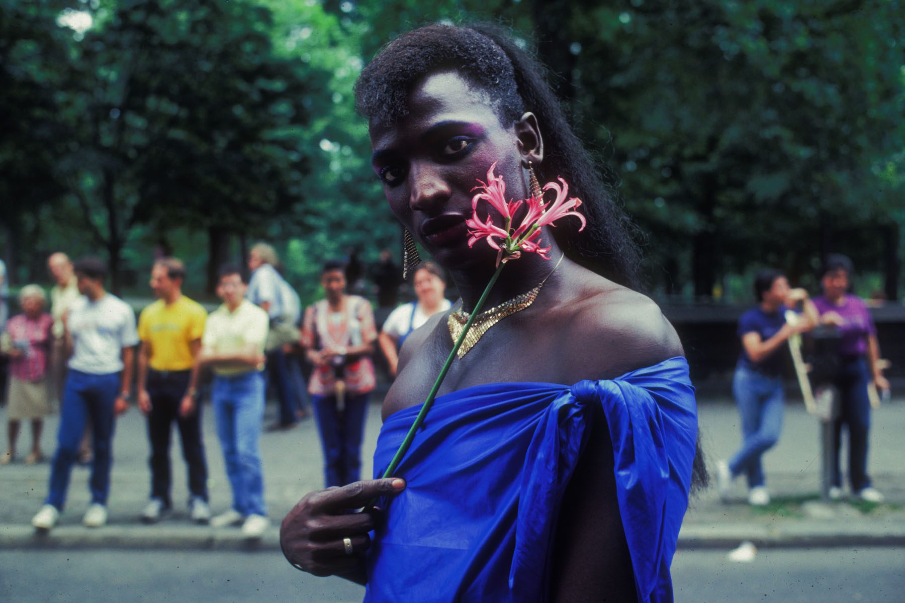 A participant wearing a golden necklace, earrings and a blue fabric as a top holds a flower to their cheek.