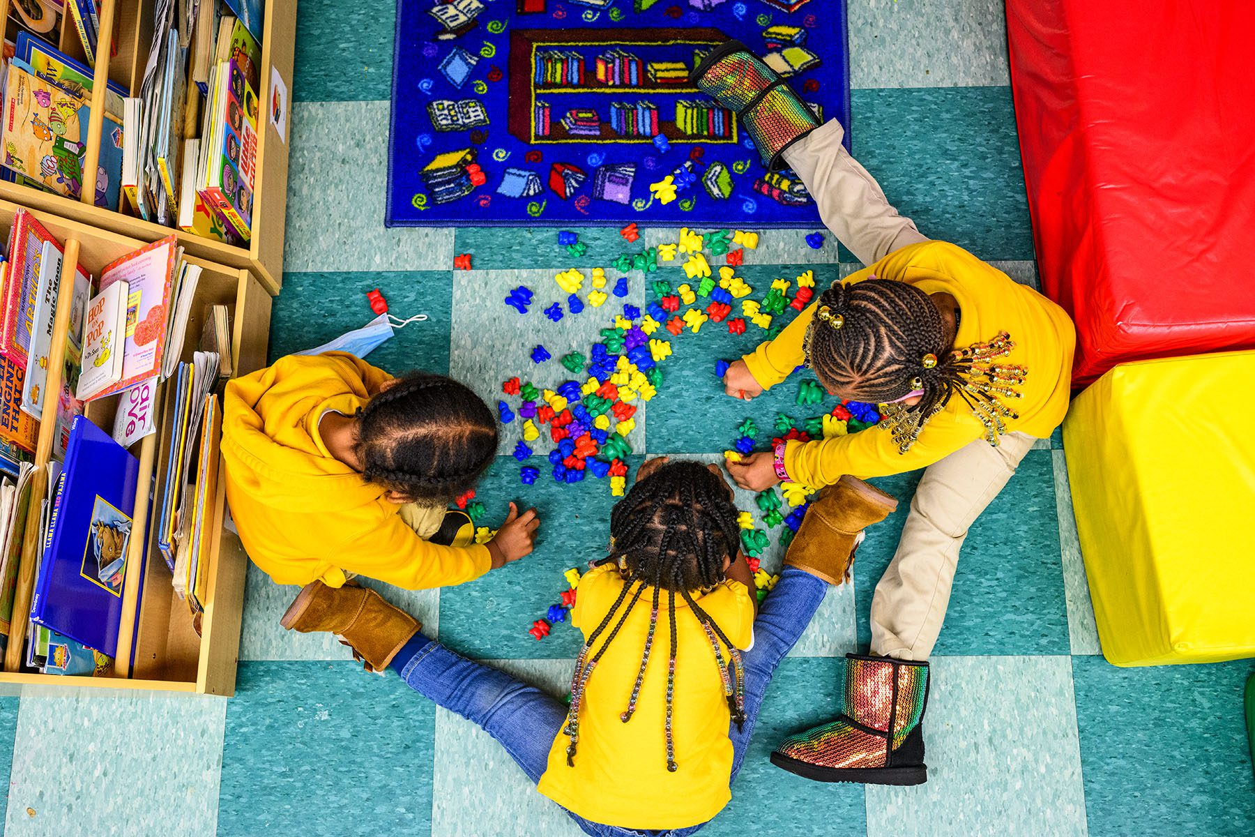 Three little girls seen from above play on a colorful classroom floor.