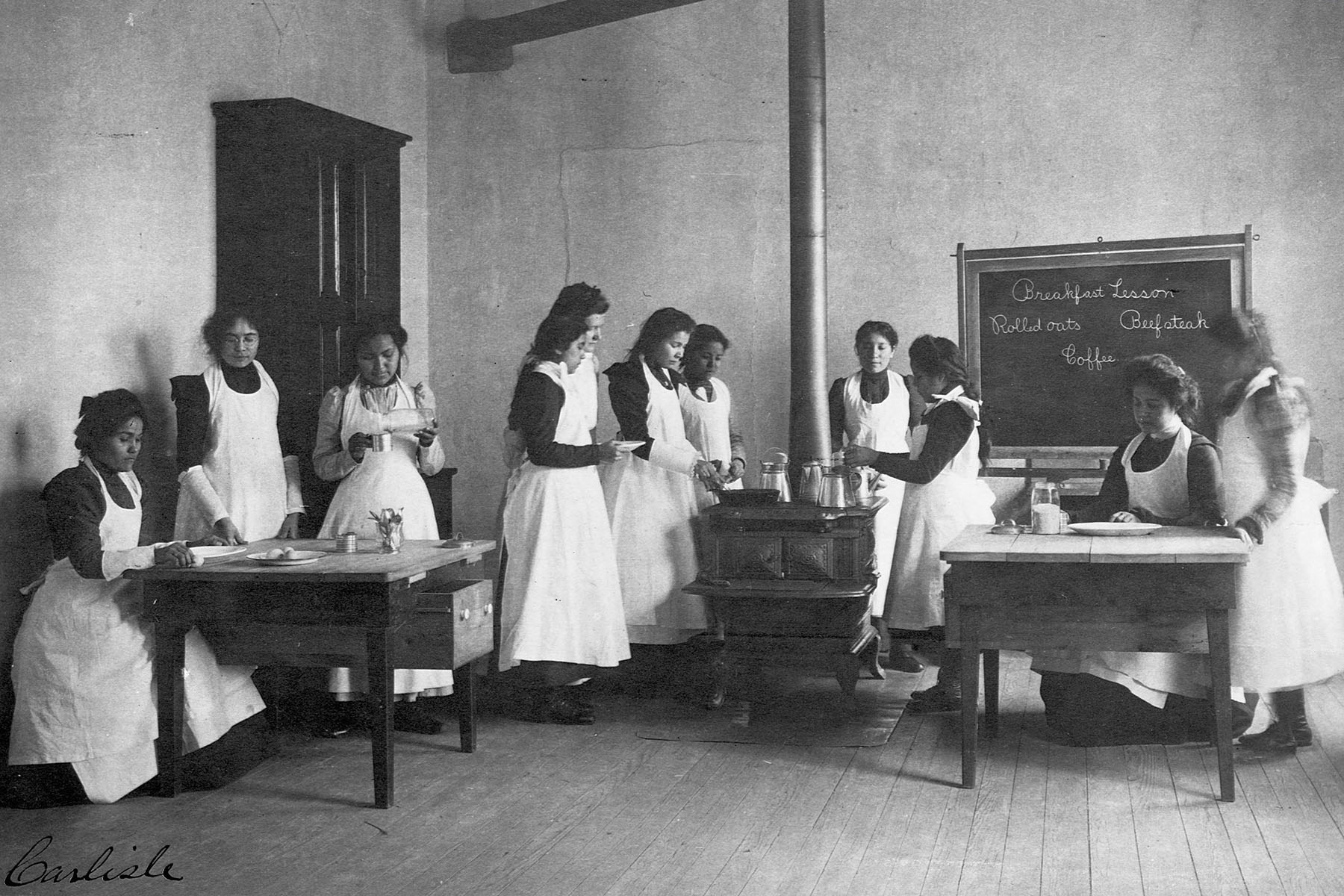 Native American girls learn to cook breakfast on wood burning stoves in a classroom.