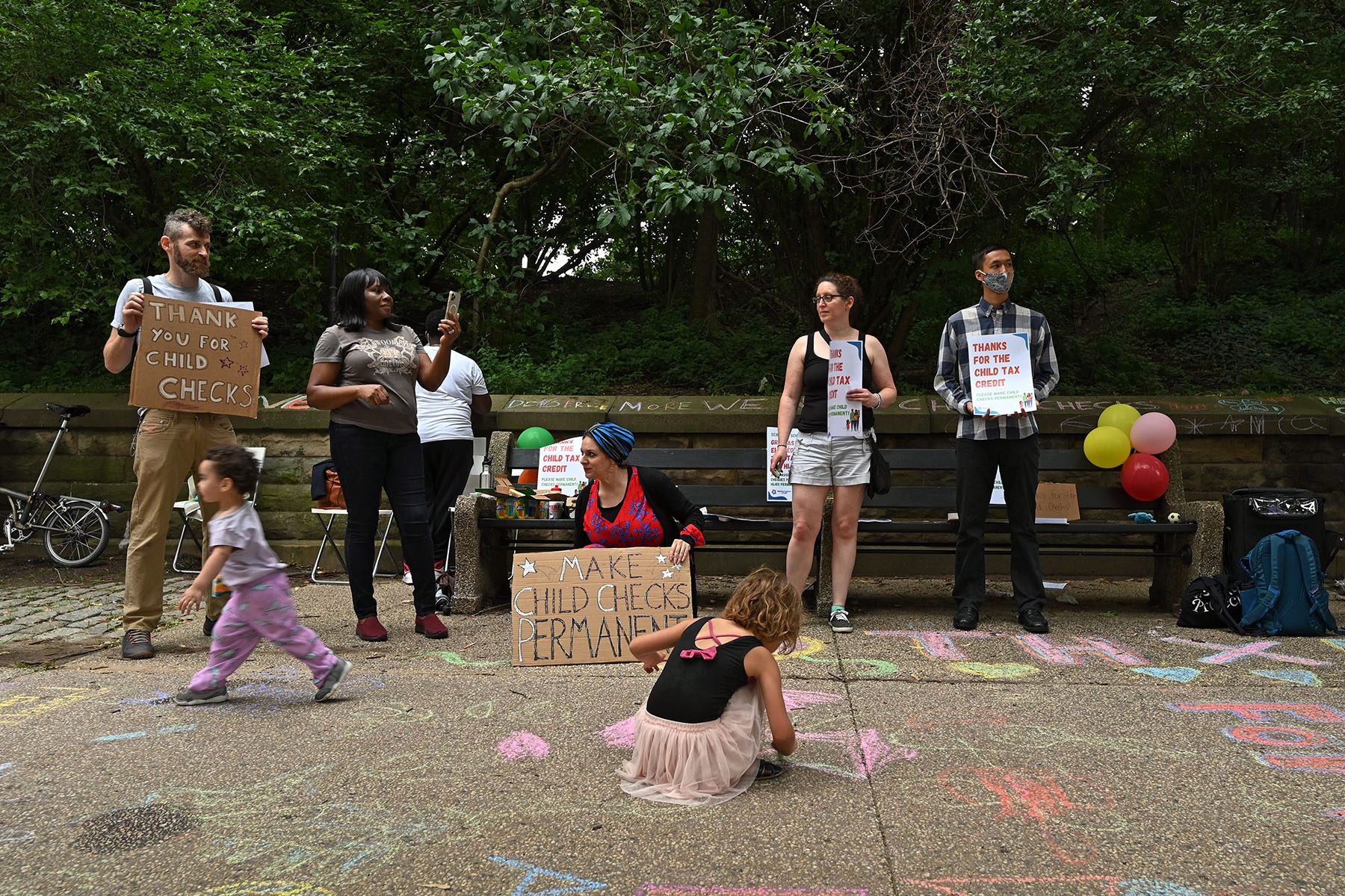 Kids draw on the sidewalk using chalk while parents hold signs.