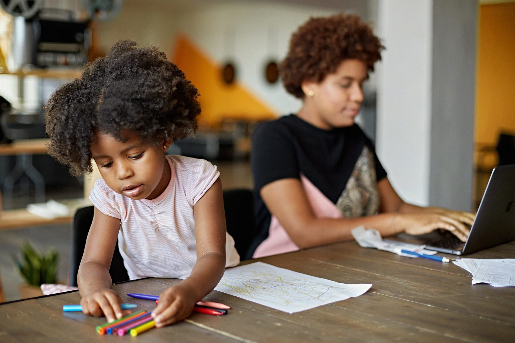 A child and woman sit together at a table while the child colors and the mother works on a laptop.