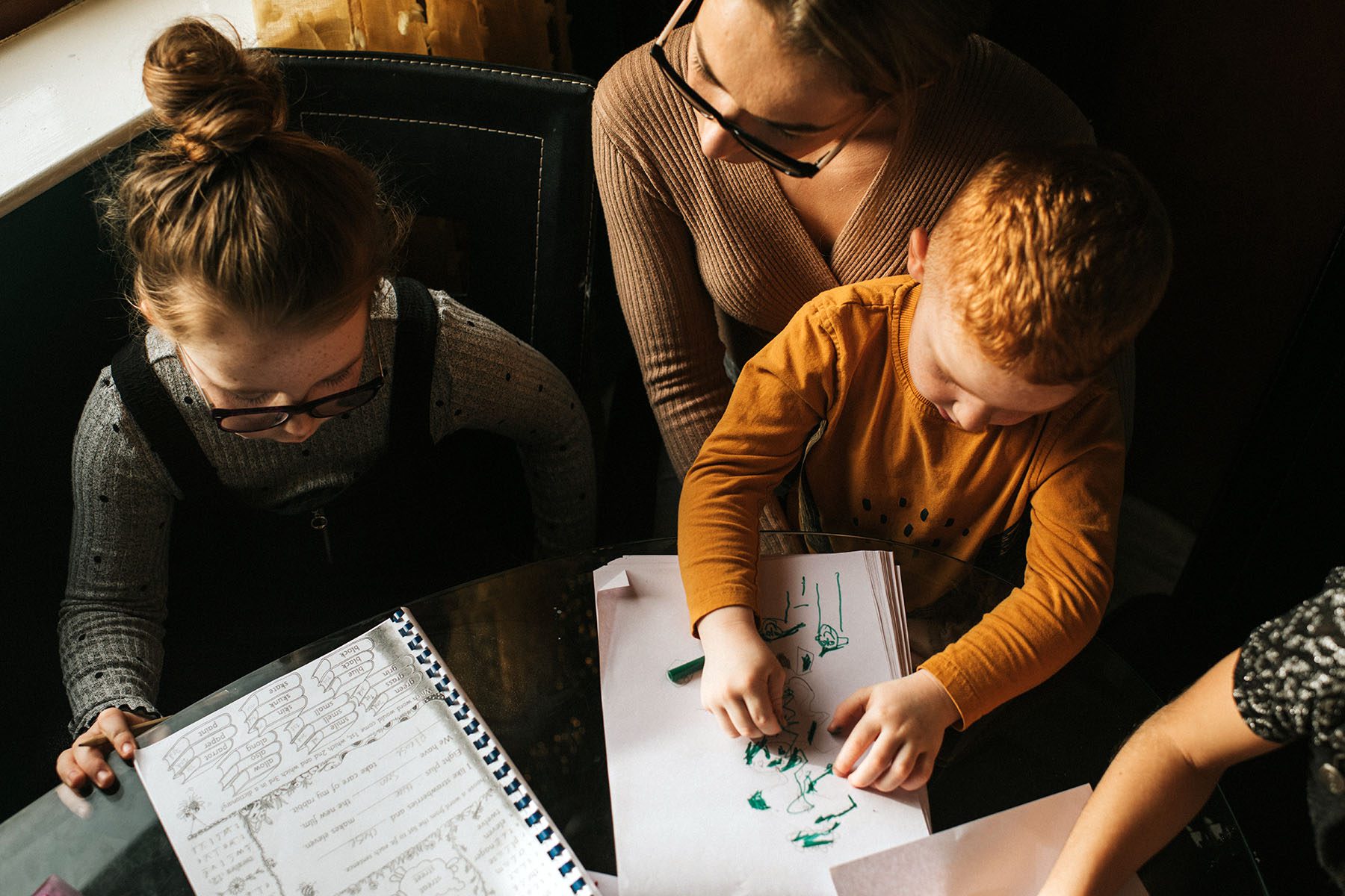 Little boy sits on his Mom's lap and draws while she helps older daughter with her schoolwork.