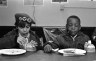 Two young boys seated at a table eat breakfast. The boy on the left wears a beret with buttons that read "Free Huey," "Boycott Grapes," and "Free Bobby."