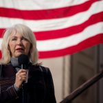 Tina Peters speaks into a microphone. An American flag can be seen in the background.