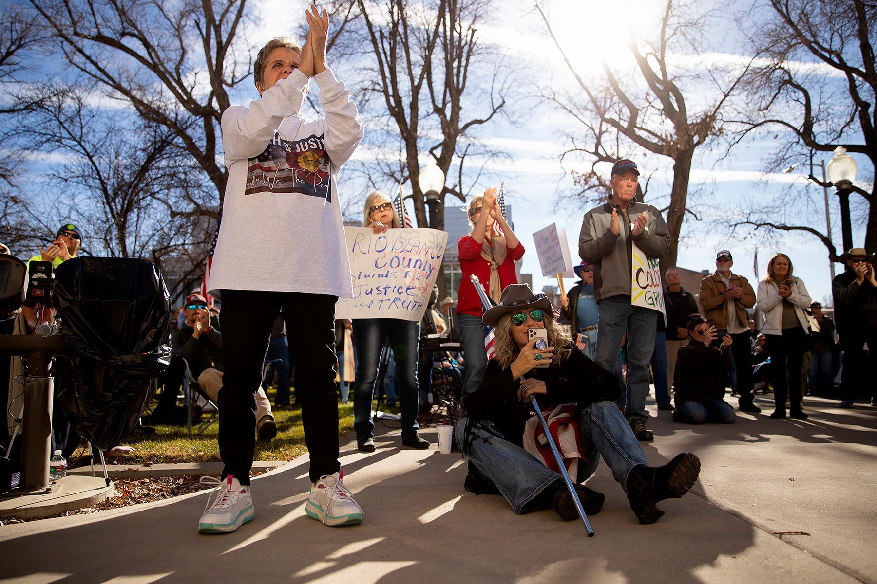 People gather at rally in support of Tina Peters. Several people clap or hold signs. One sign reads "Rio Blanco County stands for Justice & Truth."