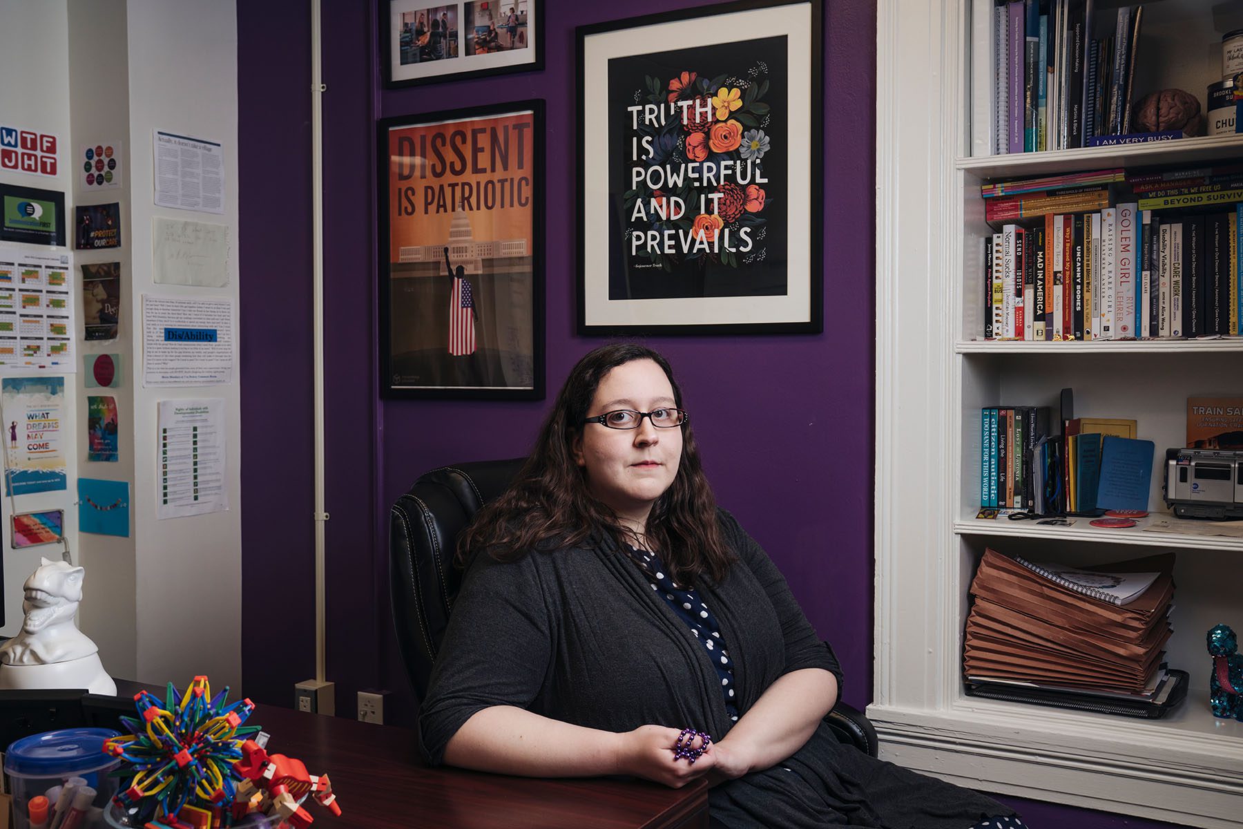 Julia Bascom poses in her DC office. There are pictures on the wall and knickknacks on the desk. Behind her two posters read "Truth is Powerful and Prevails" and "Dissent is Patriotic."