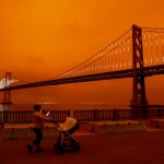 A woman pushing a stroller is seen near the Bay Bridge as a dark orange sky hands over downtown San Francisco.
