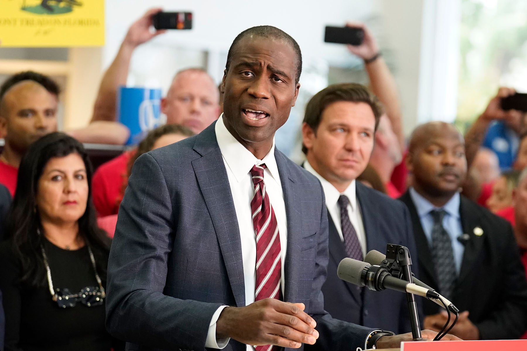 Dr. Joseph A. Ladapo is seen speaking at a podium. He is surrounded by people including Florida governor Ron DeSantis, who are blurred in the background.