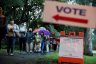 Voters wait in line to cast their ballots on a rainy florida morning. Several people in line are holding umbrellas and wearing face masks. A 