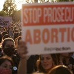A large crowd of people protest in front of the Supreme Court. A sign in the foreground reads 