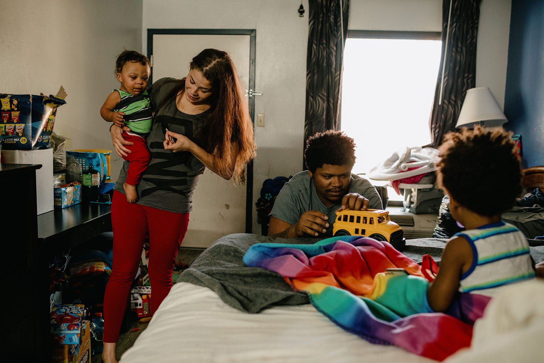 A mother smiles as she holds her baby and a dad plays with a toy car as his son looks on in a small hotel room.