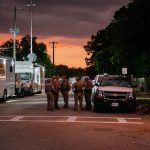 Law enforcement officers speak together outside of Robb Elementary School in the evening. The colors of the sunset are reflected on the street.