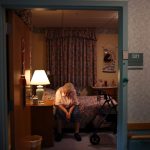 A woman sits on a bed in a dark room in a nursing home.