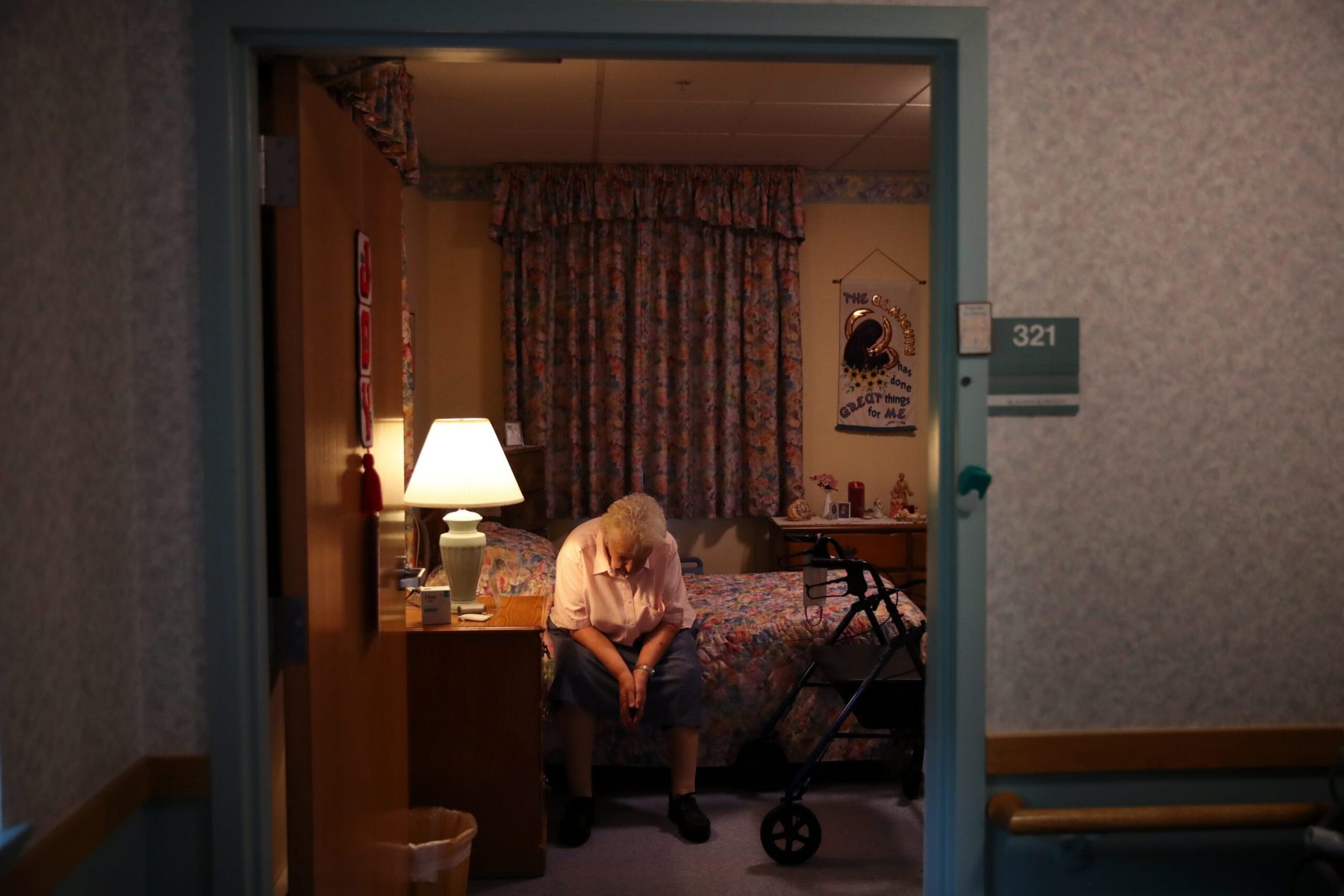 A woman sits on a bed in a dark room in a nursing home.