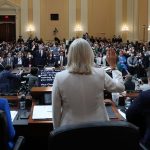 View of committee hearing room, from behind members, with Liz Cheney standing to deliver the oath