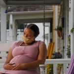 A pregnant woman wearing a face mask sits on a swing on her front porch.