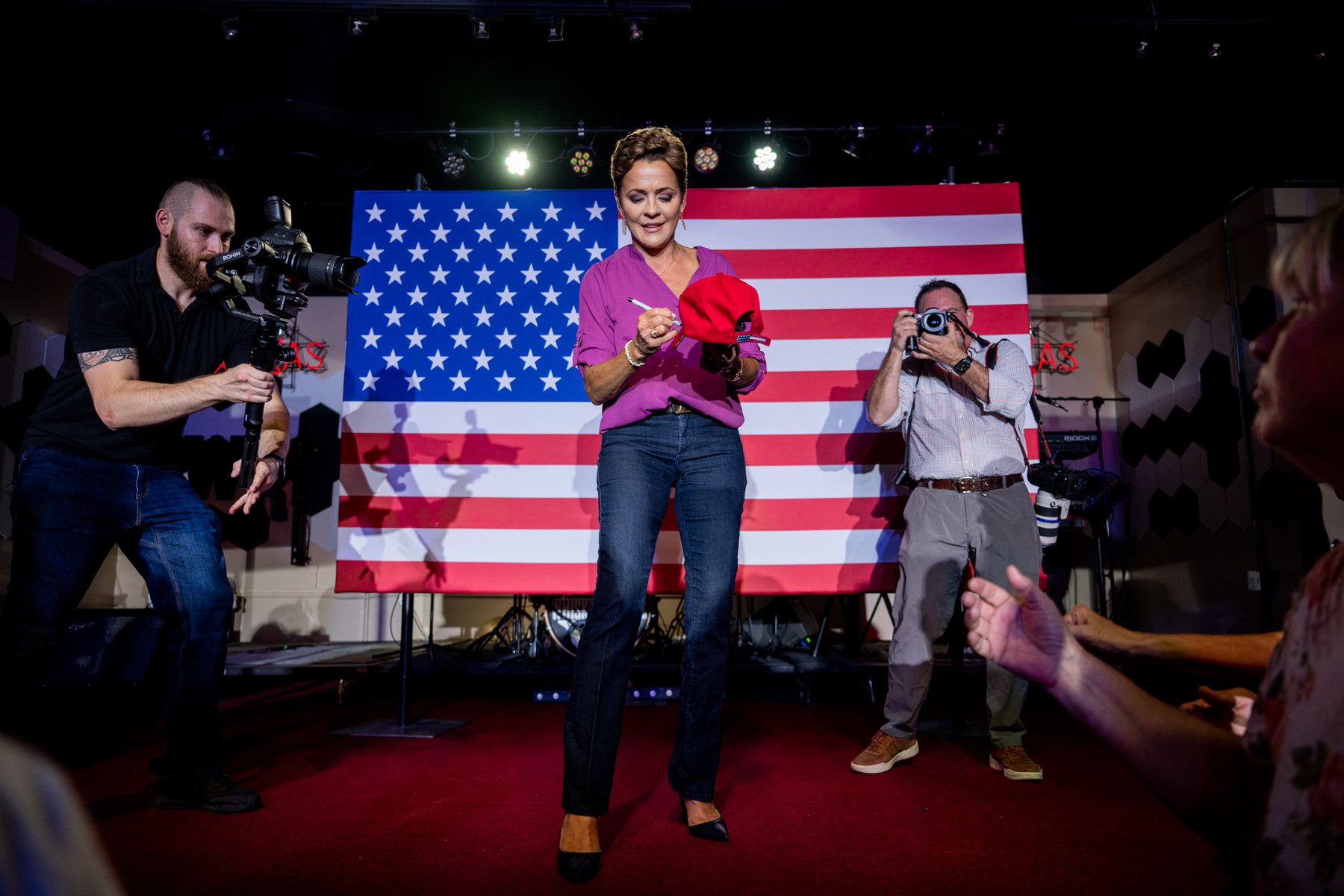 Kari Lake stands in front of an American flag and signs a hat