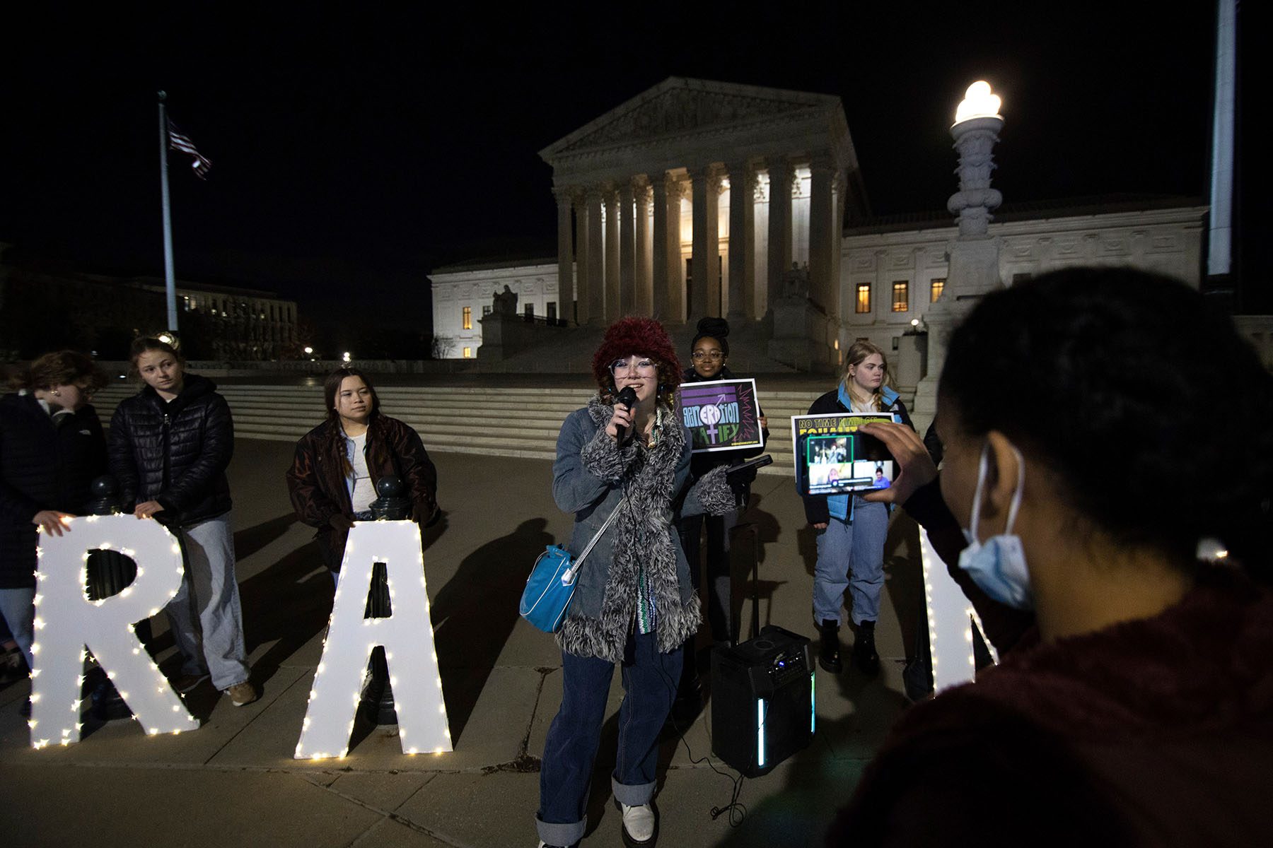 A member of generation ratify speaks into a microphone as another live streams the rally.