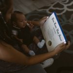 A woman holding a baby holds her high school diploma.