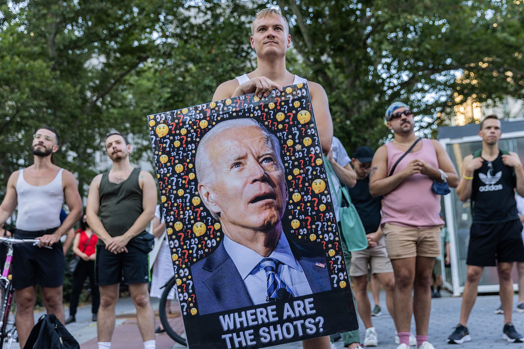 A protester holds a large image of President Biden looking bewildered and surrounded by question mark and thinking face emojis. Under the image are the words "Where are the shots."