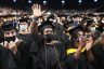 A graduate, surrounded by other graduates in caps and gowns, waives during a commencement ceremony.