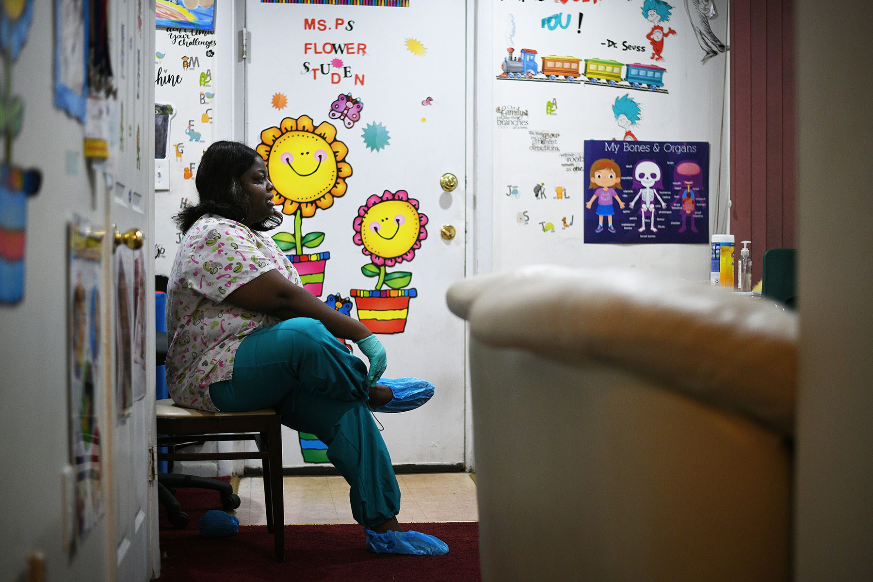 A woman puts on protective gloves and slippers before starting work at her daycare center inside her home. The walls are decorated with Dr. Seuss quotes and characters and informative posters.