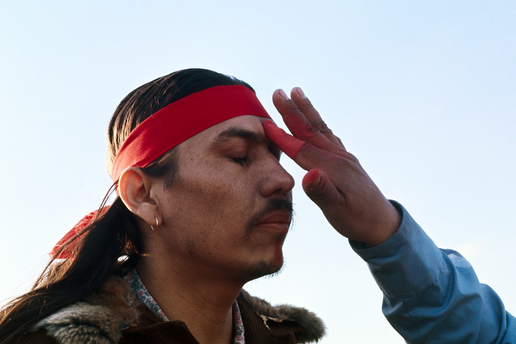 A medicine man applies red war paint to a warrior's forehead as the warrior closes his eyes.