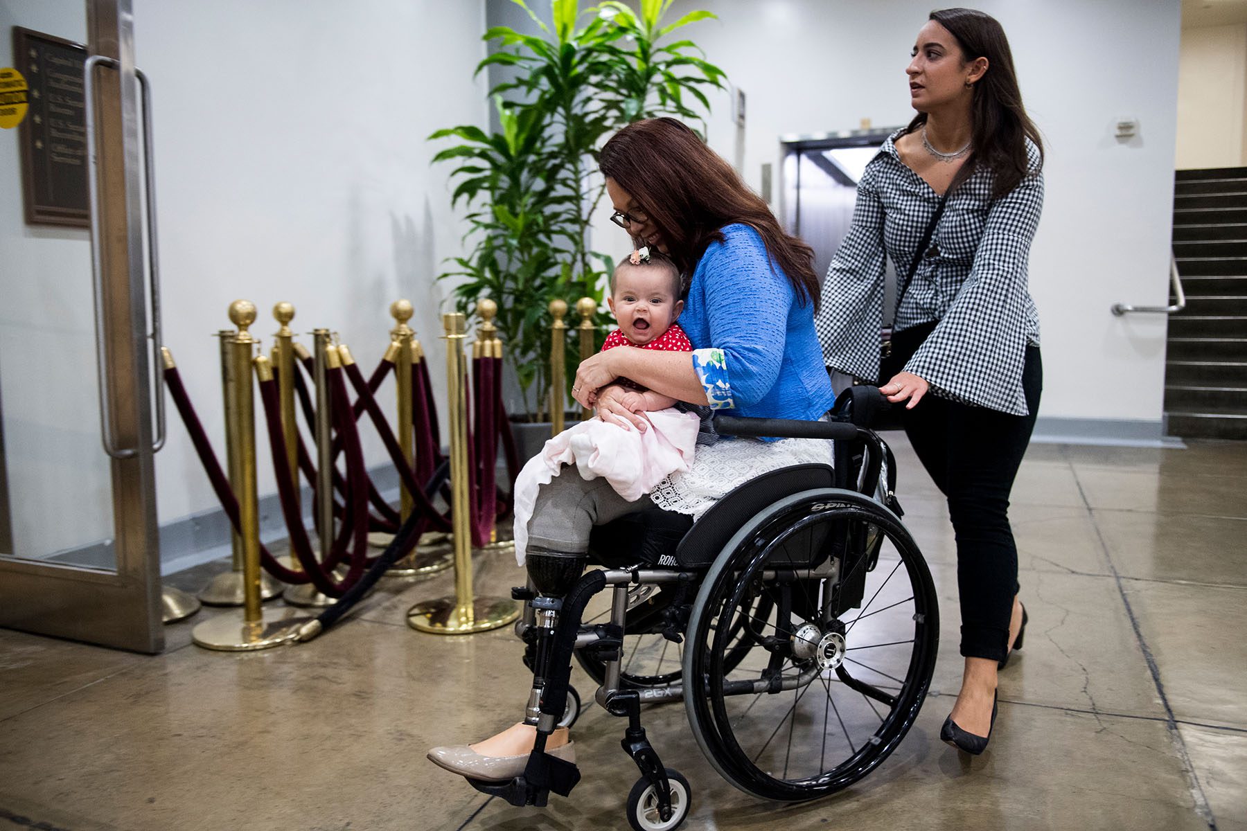 Sen. Tammy Duckworth holds her daughter Maile as they both smile. A woman pushes Sen. Duckworth's wheelchair.